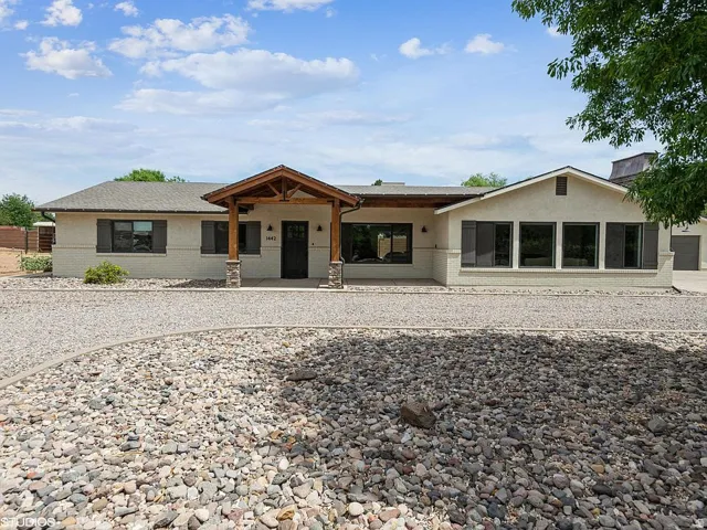 Stucco exterior with a pitched roof, front entry porch with wood columns and stone accents, multiple large windows, and a gravel driveway