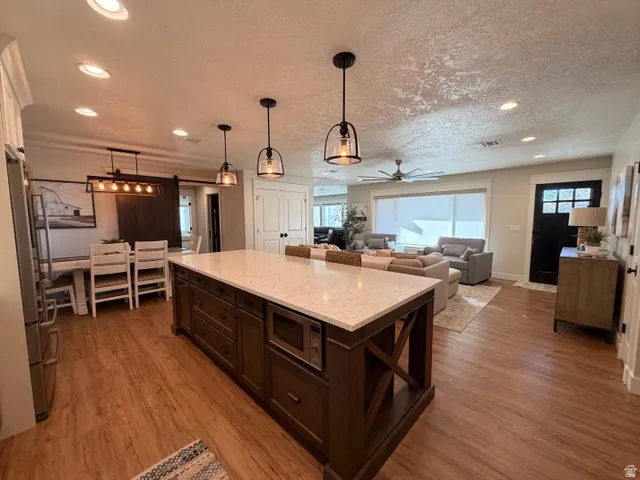 Expansive kitchen island with a light-toned countertop, wood-finish flooring throughout, recessed ceiling lighting, and pendant light fixtures
