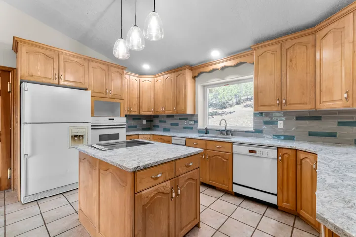 Kitchen featuring light wood cabinetry, white appliances, a center island with a cooktop, speckled countertops, and a tiled backsplash