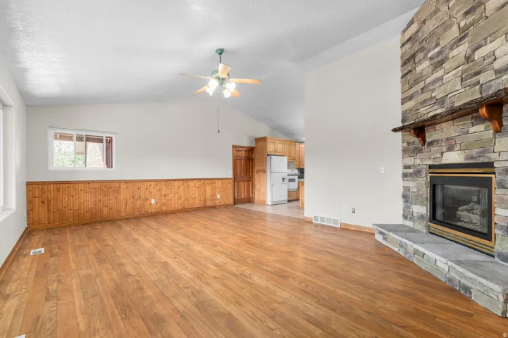 Vaulted ceiling with a ceiling fan, stone-clad fireplace with a wood mantle, wood-finish flooring, and wood wainscoting
