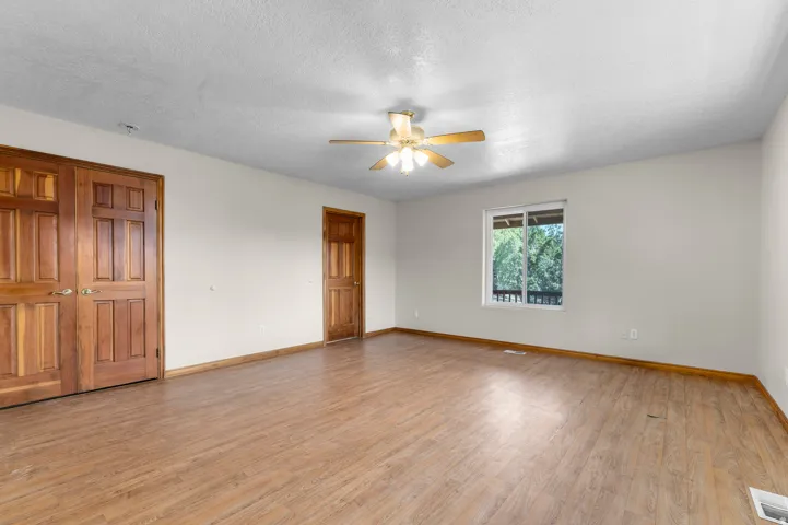 Wood-finish flooring throughout, two panel doors, a ceiling fan with integrated lighting, and a window overlooking exterior greenery