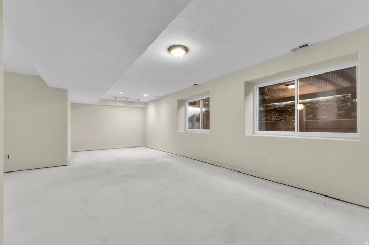 Spacious room with light-colored walls and flooring, featuring two large windows, a ceiling-mounted light fixture, and recessed lighting