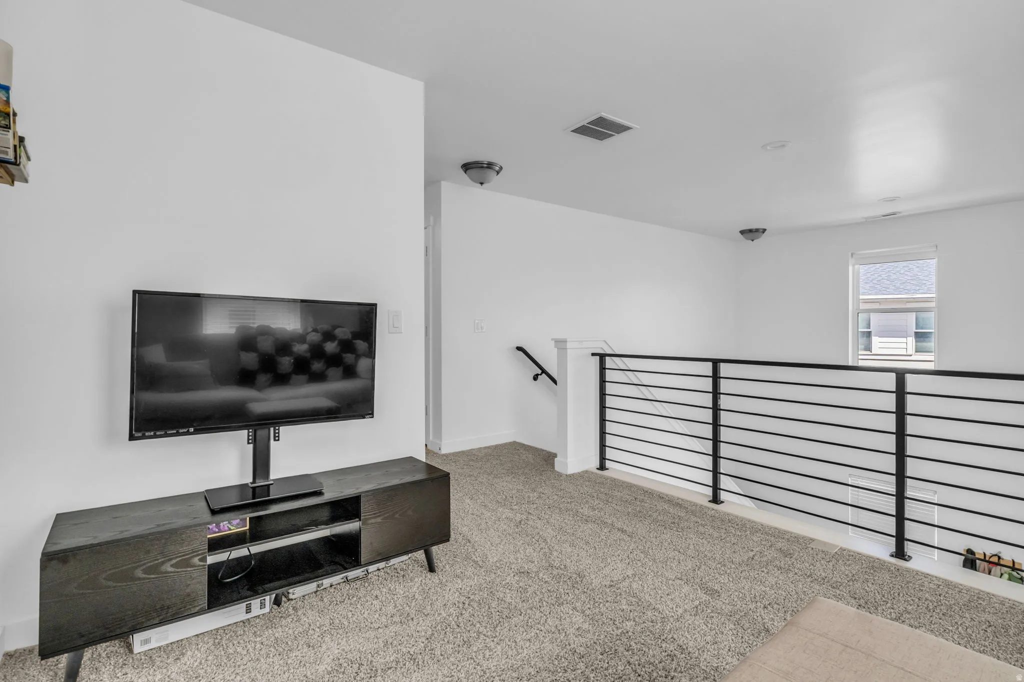 Carpeted upper-level space featuring a contemporary horizontal metal railing, a single window, and a wall-mounted light fixture