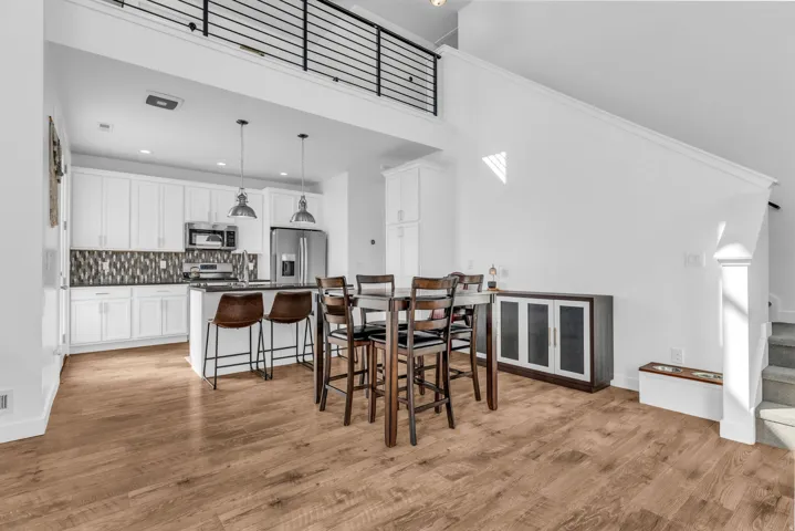 Open-concept living area featuring wood-finish flooring, a contemporary kitchen with white cabinetry, a kitchen island with pendant lighting, and a staircase with a metal railing