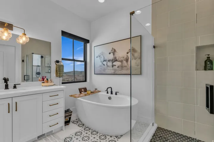 Freestanding soaking tub with a black wall-mounted faucet, dual vanity with white cabinetry and a white countertop, walk-in shower with a glass enclosure, and patterned tile flooring