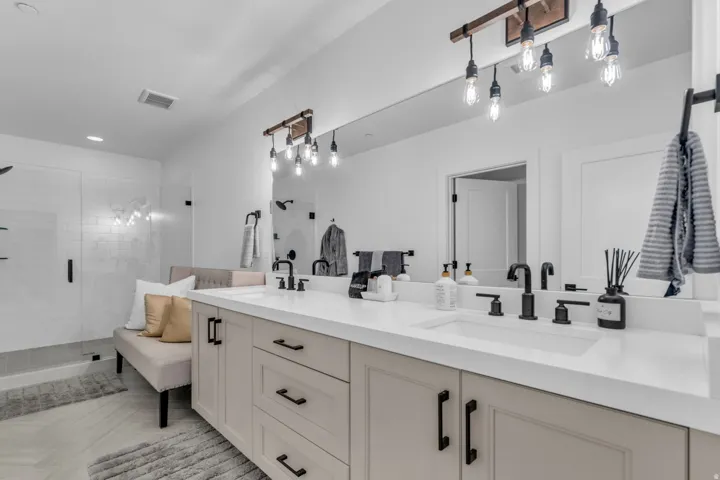 Double vanity with integrated sinks and white countertops, matte black hardware, exposed bulb light fixtures, and a glass-enclosed shower featuring subway tile