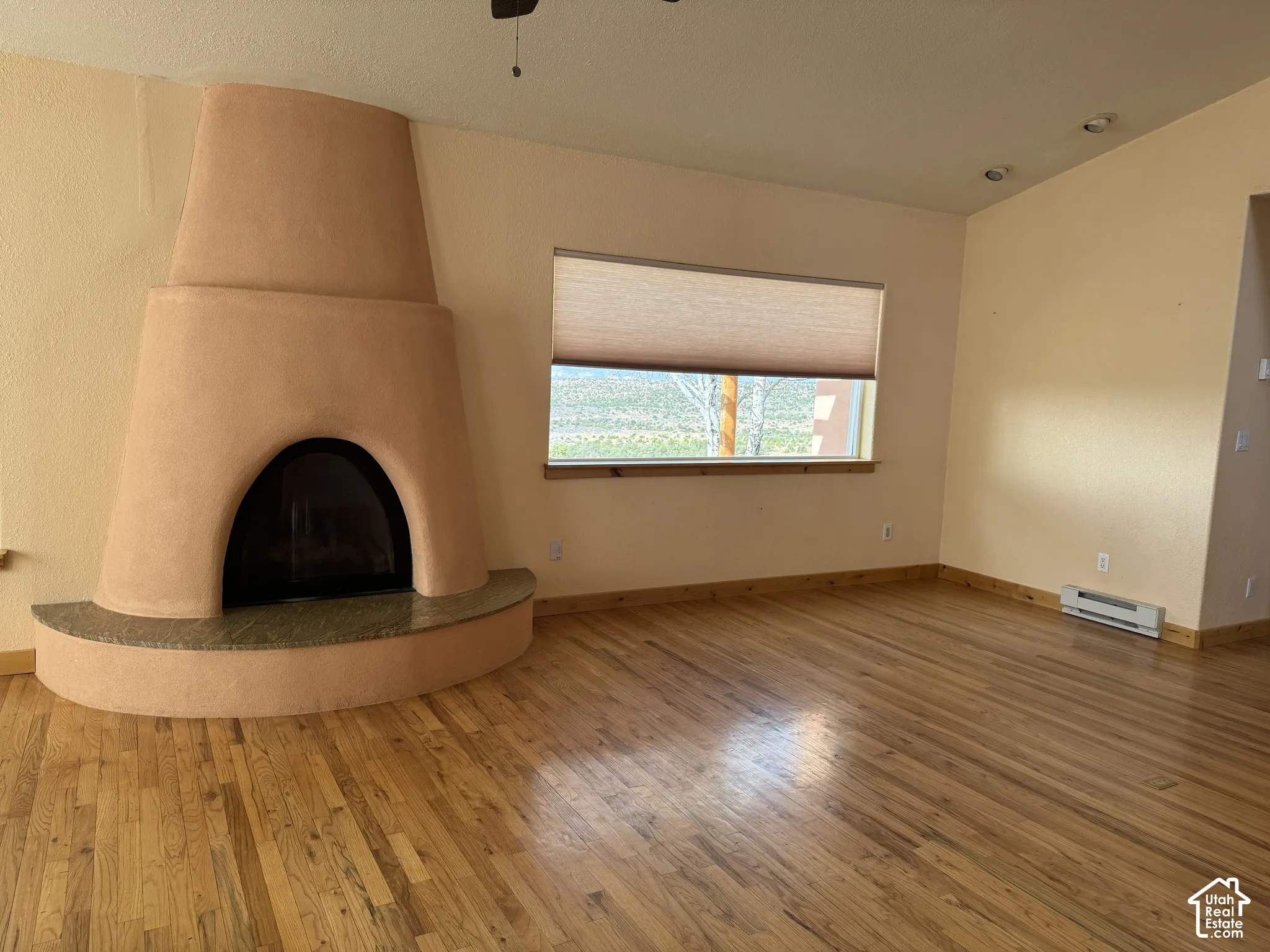 Kiva fireplace with a stone hearth, wood-finish flooring, recessed lighting, and a window with roller shade
