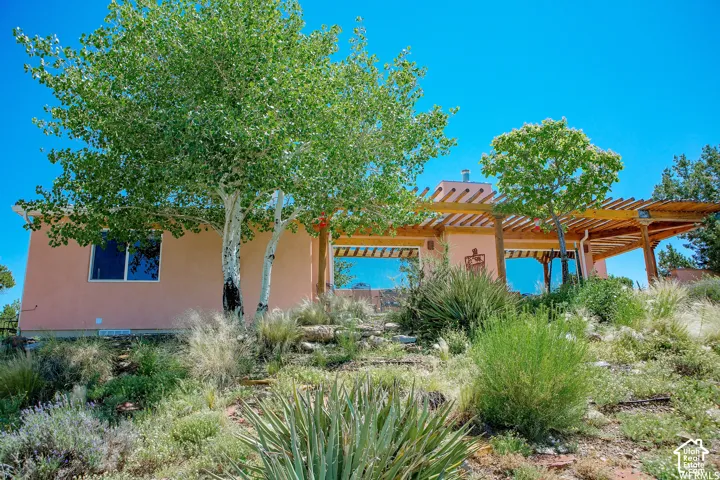 Stucco exterior with a wood pergola, multiple large windows, and a sloped, landscaped lot featuring mature trees and native plantings