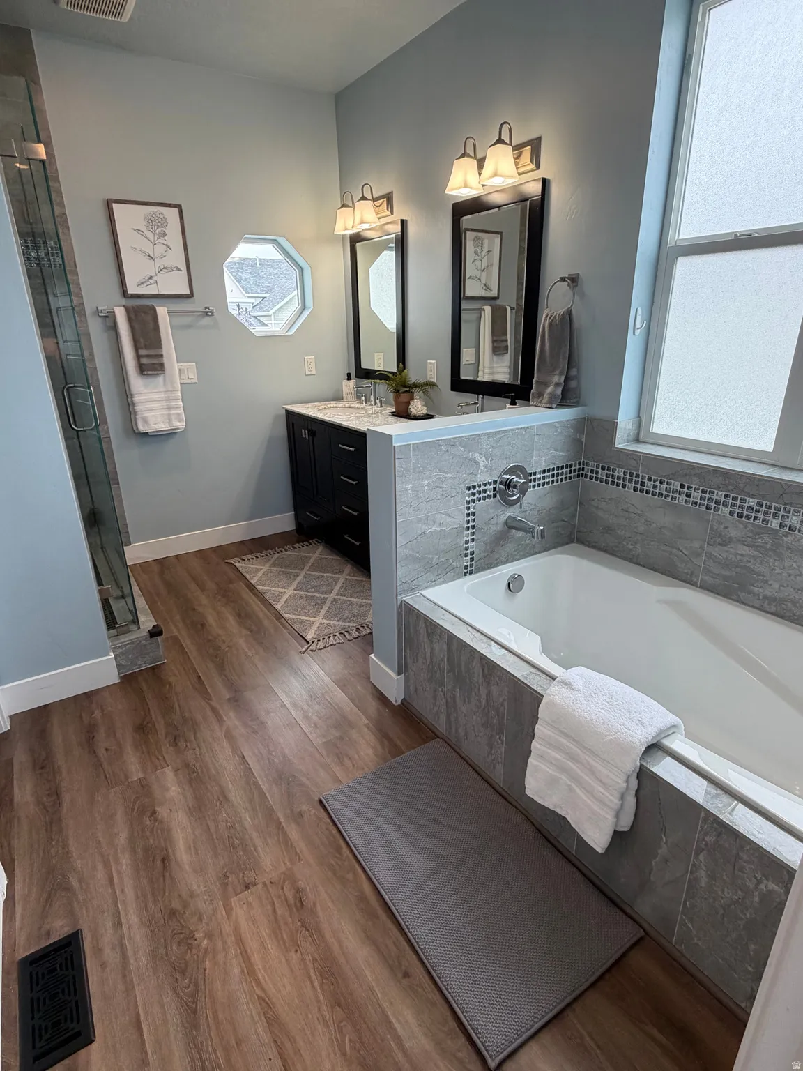 Primary Bath. LVPflooring, built-in soaking tub with tiled surround, glass-enclosed shower, dual vanity with undermount sinks, and wall-mounted light fixtures