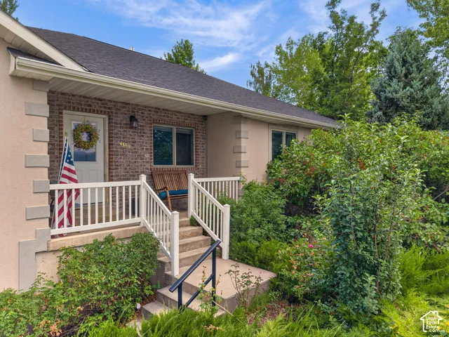 Brick facade, covered front porch, white railing, concrete steps, and mature landscaping
