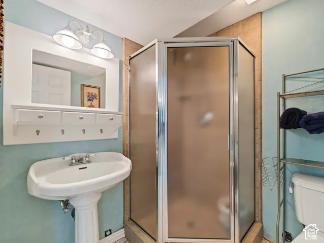 Basement bathroom with pedestal sink, chrome faucet, wall-mounted mirror with integrated lighting, built-in shelving with hooks, and a corner shower with frosted glass enclosure
