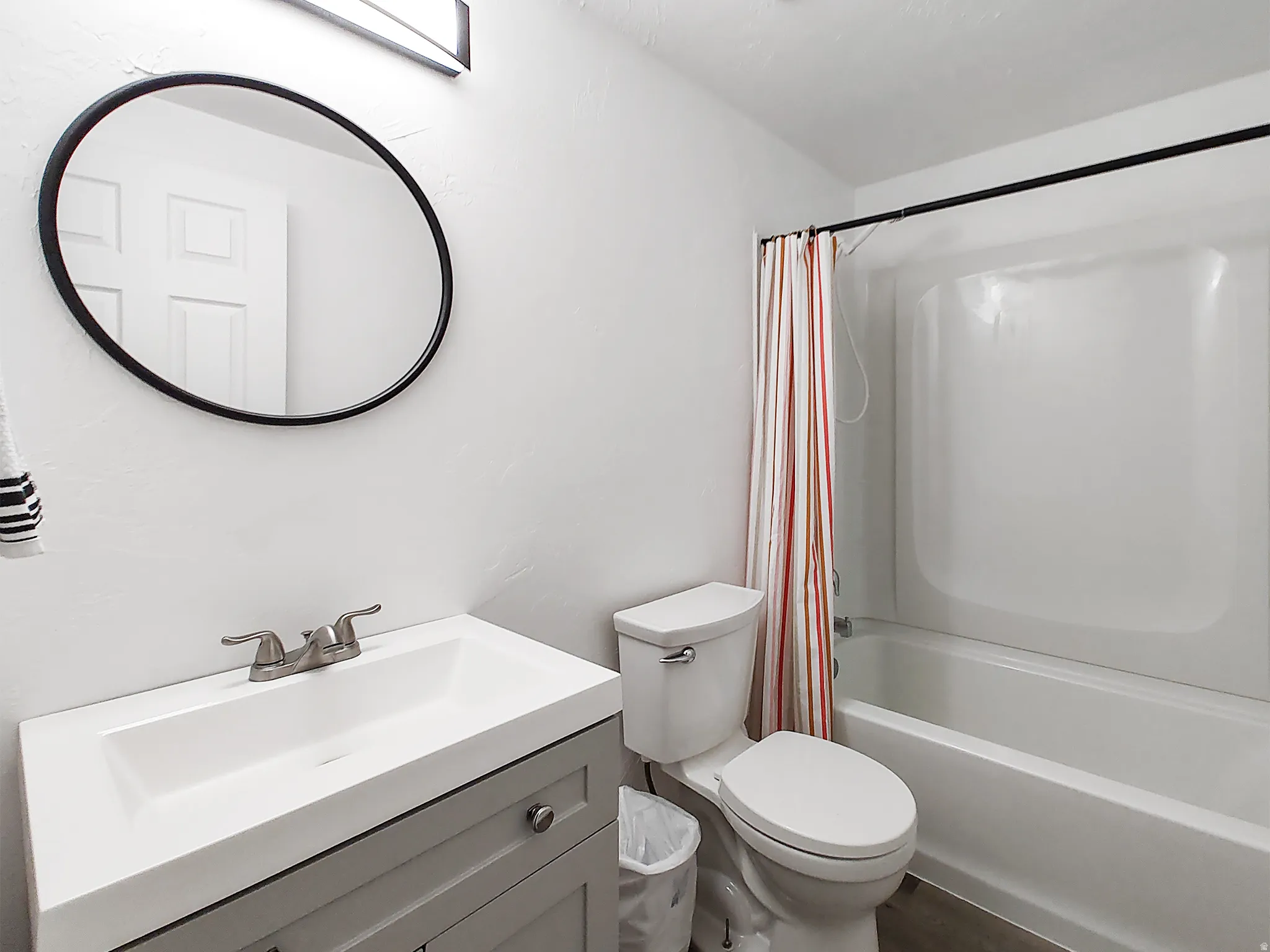 White walls, a white integrated sink, a gray vanity cabinet with silver hardware, a black-framed circular mirror, and a white bathtub with a shower