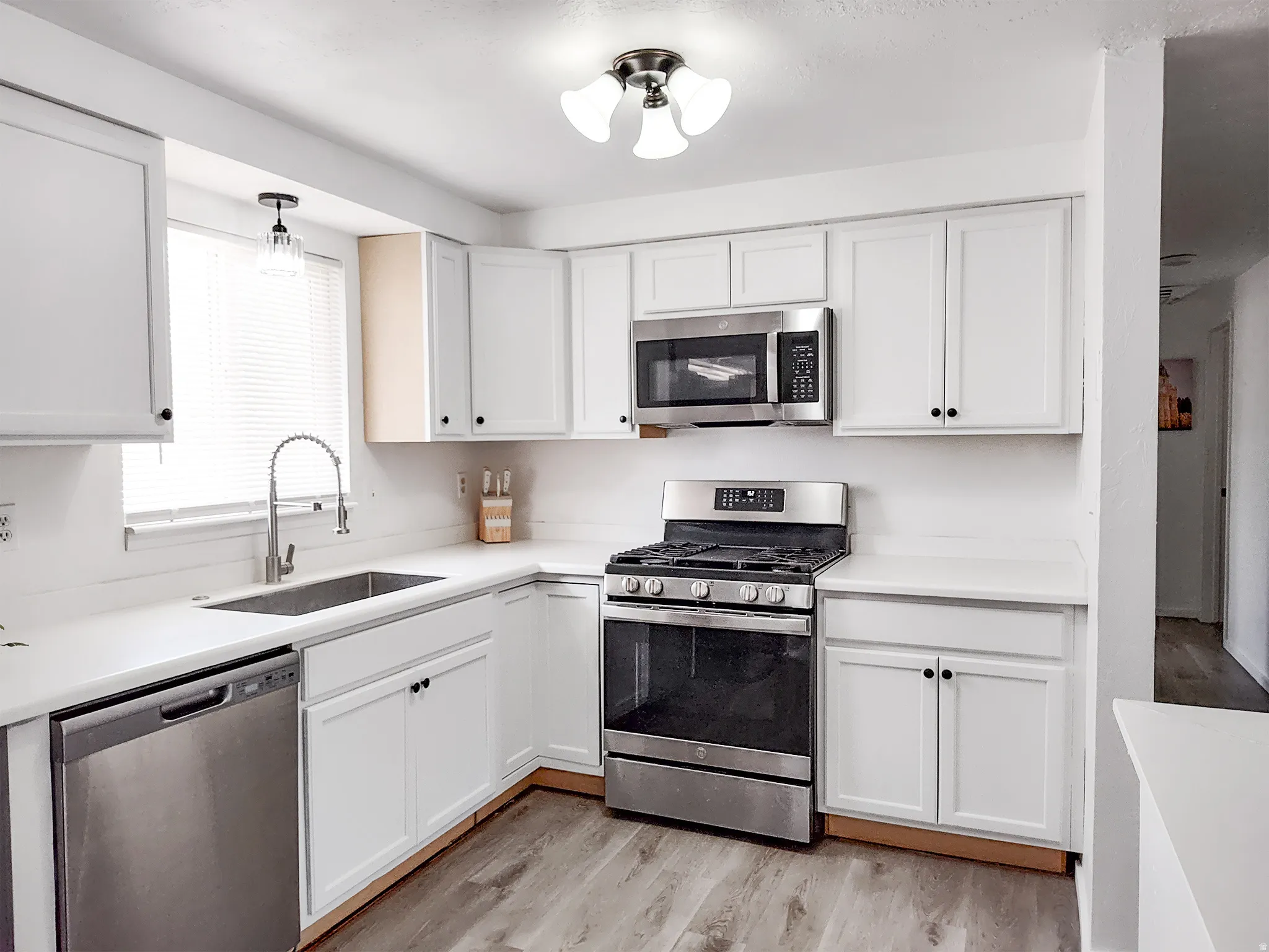 White shaker-style cabinetry, stainless steel appliances, light-toned countertops, wood-finish flooring, and a single-basin sink with a pull-down faucet