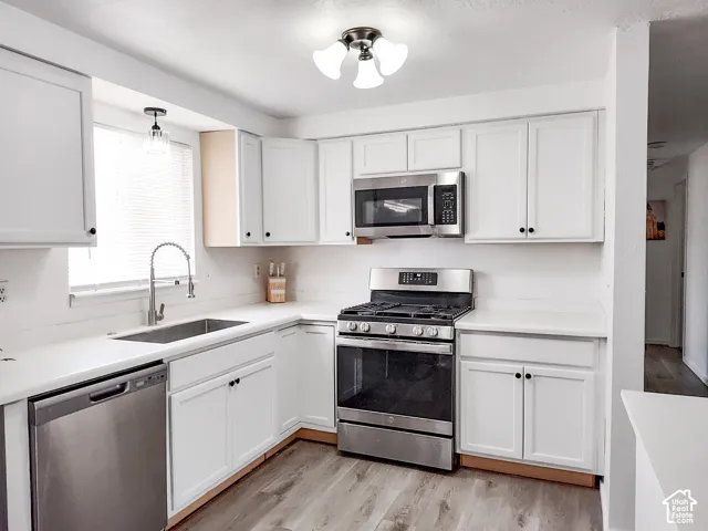 White shaker-style cabinetry, stainless steel appliances, light-toned countertops, wood-finish flooring, and a single-basin sink with a pull-down faucet