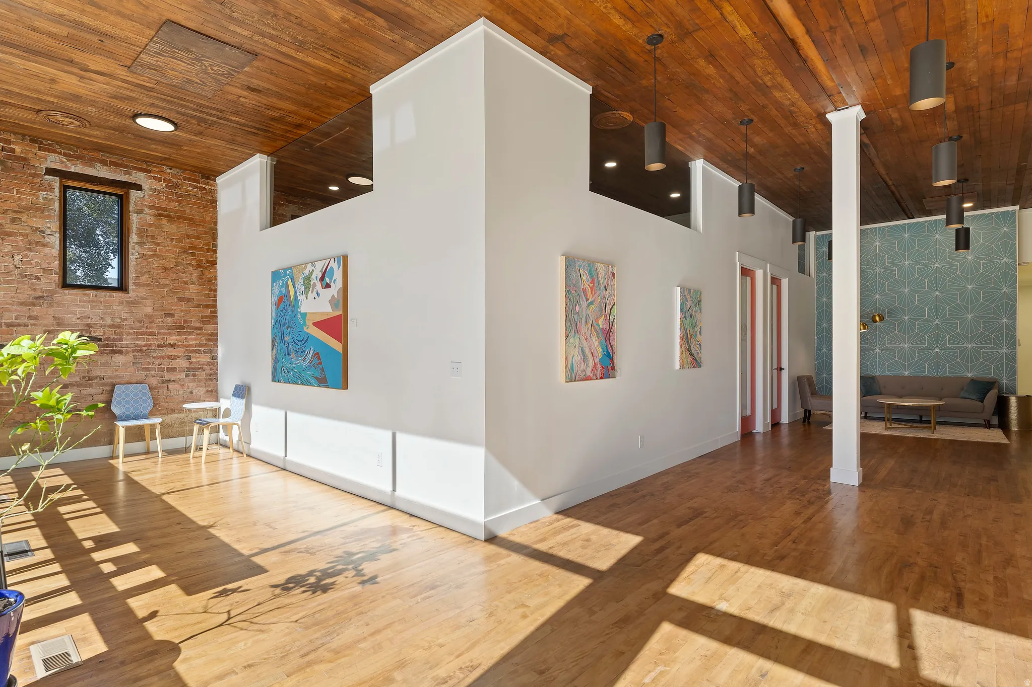 Wood-finish flooring throughout, exposed brick wall, wood-paneled ceiling, multiple pendant light fixtures, and a half-height interior wall
