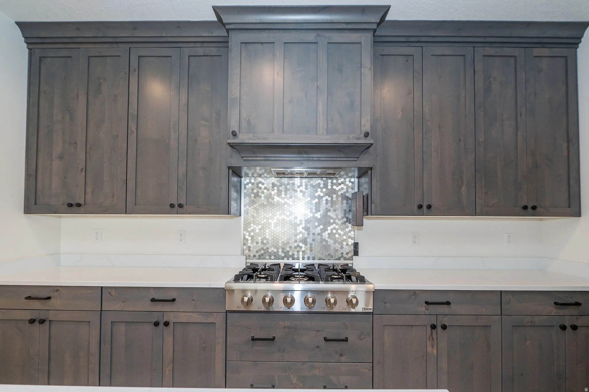 Gray wood-finish cabinetry, white countertops, built-in range hood, stainless steel gas range, and mosaic tile backsplash