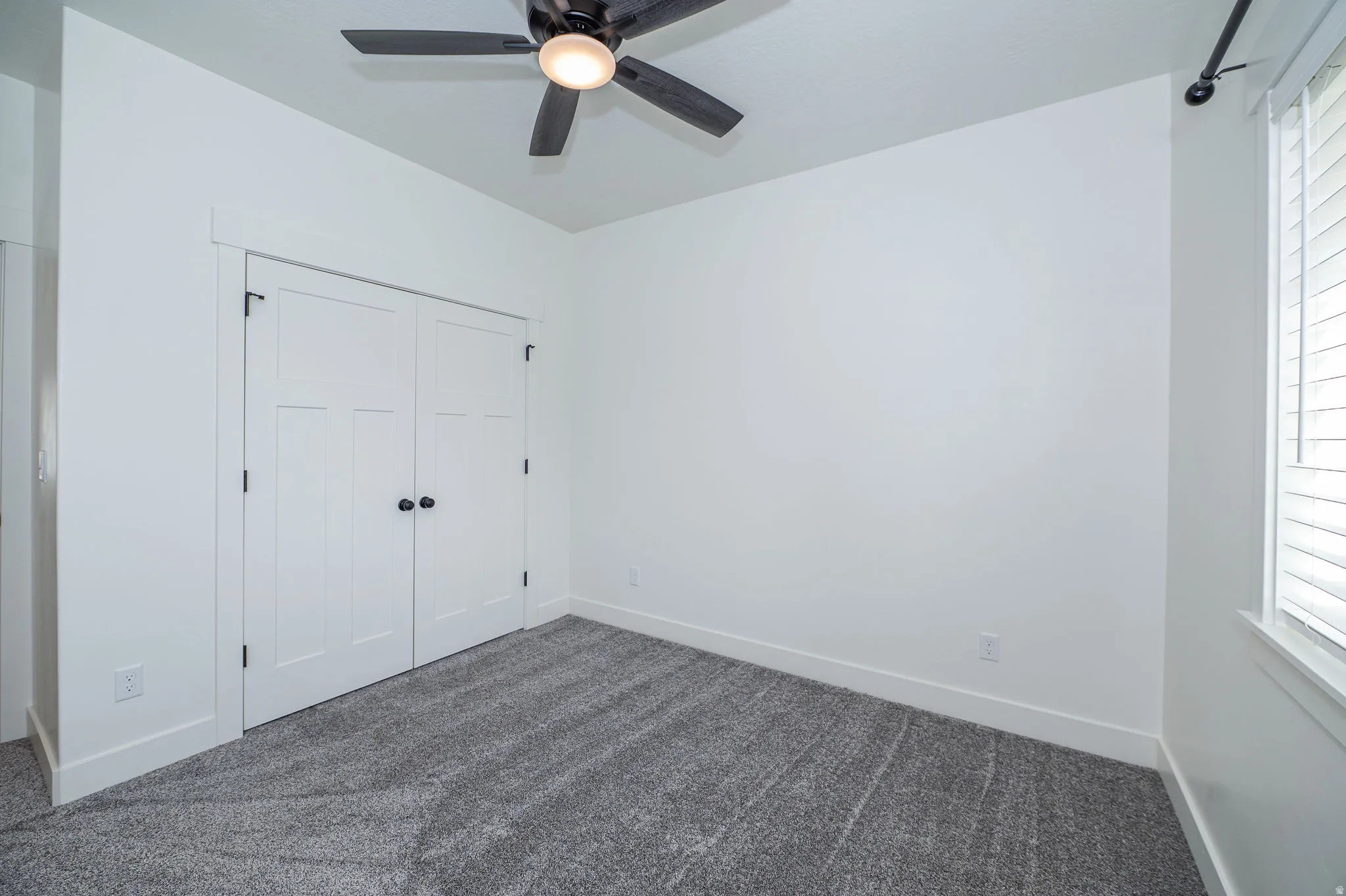 White interior walls, gray carpet flooring, white paneled bifold doors with black hardware, ceiling fan with integrated lighting, and a window with white blinds
