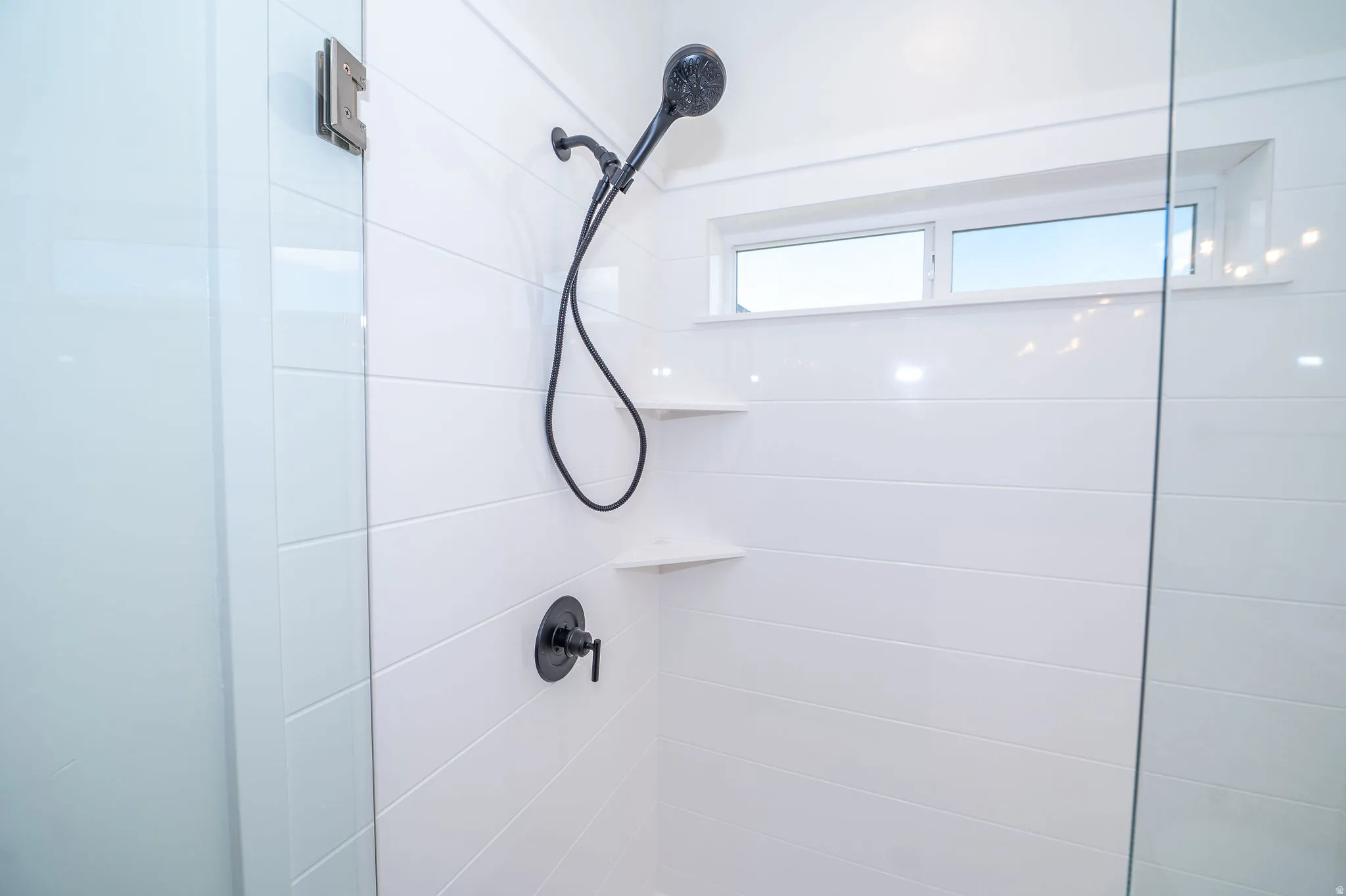 Walk-in shower with clear glass enclosure, matte black shower fixtures, two built-in corner shelves, and a horizontal window