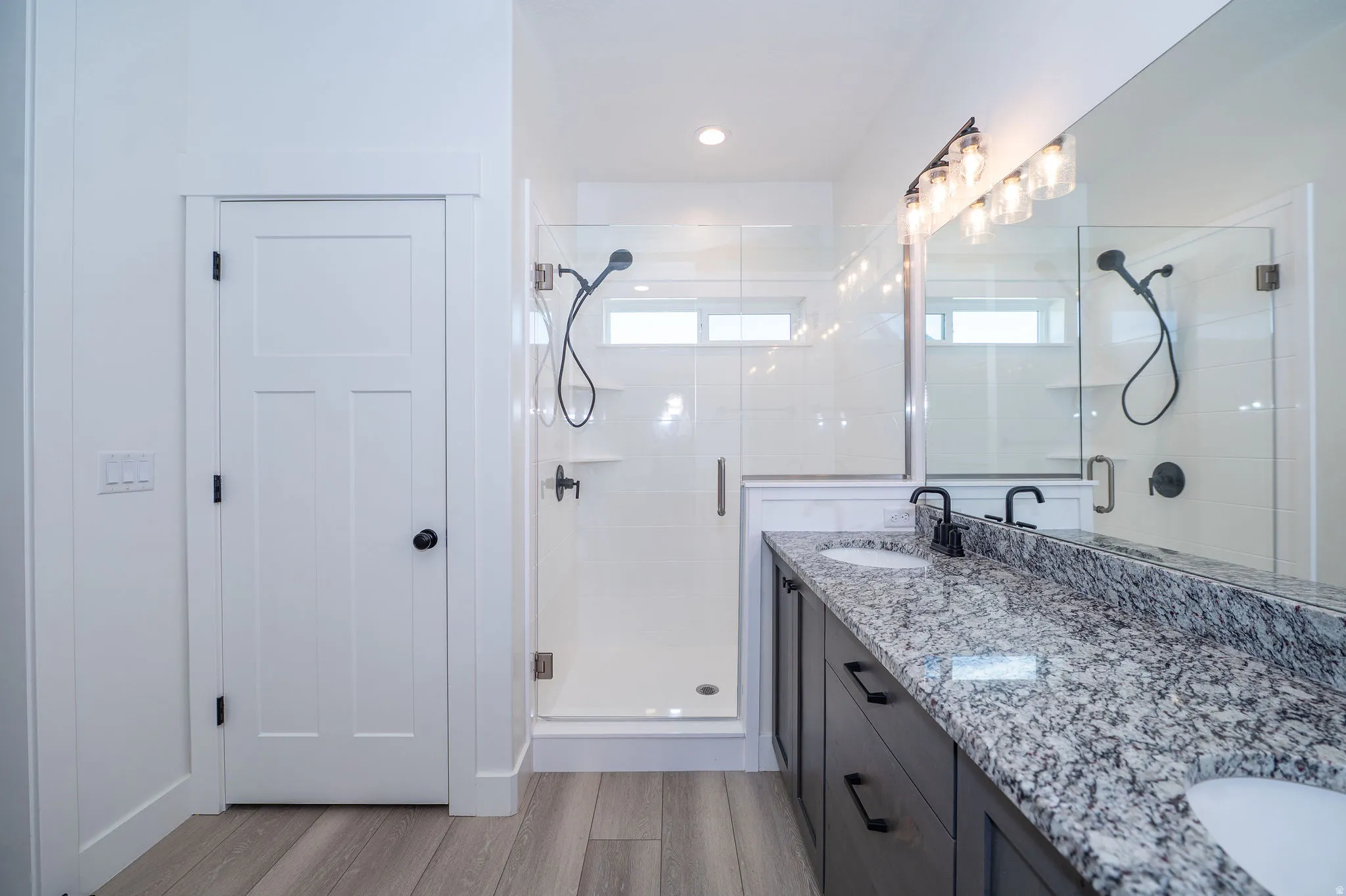 Double vanity with stone countertop, wood-finish flooring, glass-enclosed shower, and built-in shower shelving