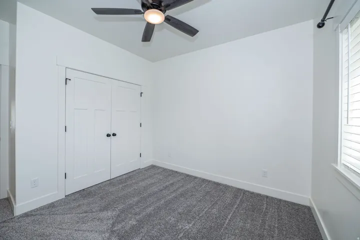 White interior walls, gray carpet flooring, white paneled bifold doors with black hardware, ceiling fan with integrated lighting, and a window with white blinds