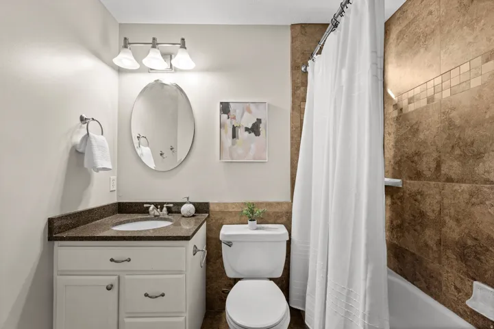 White vanity cabinet with dark countertop, oval mirror, three-light vanity fixture, and tiled shower surround with mosaic accent