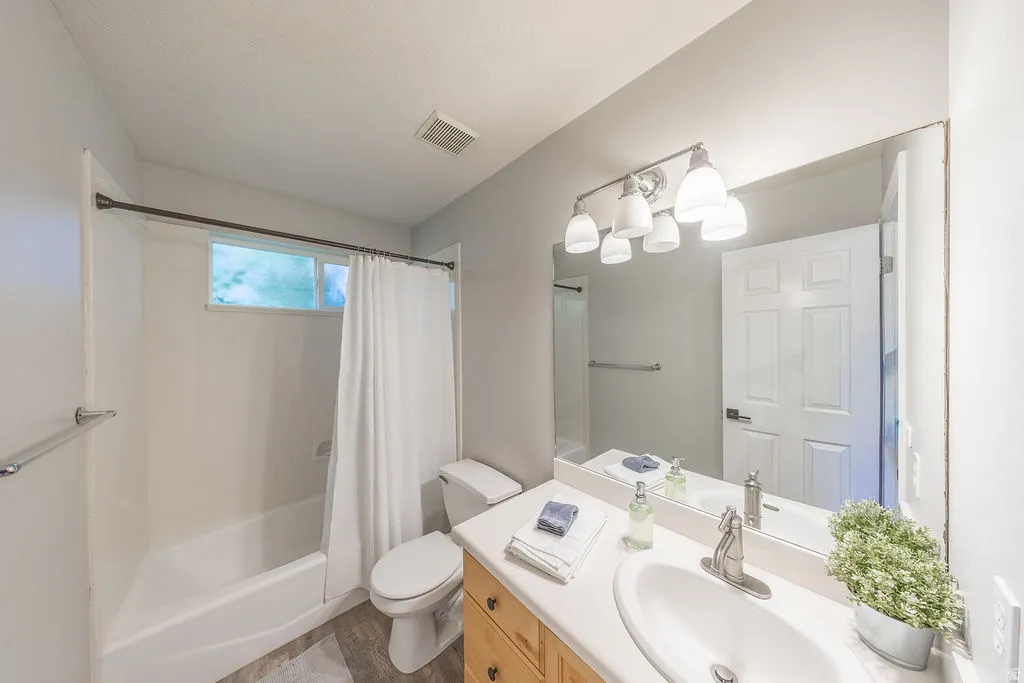 Full-width vanity mirror, four-light vanity fixture, single-basin vanity with brushed nickel faucet, wood-finish flooring, and a window above the bathtub