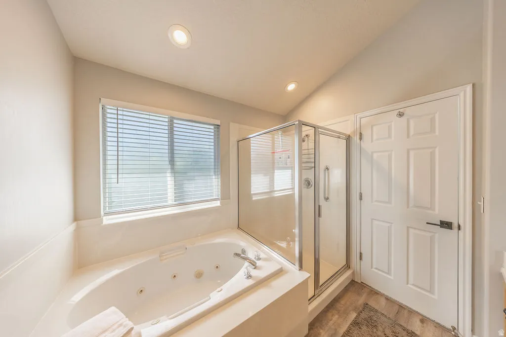 Soaking tub with integrated jets, adjacent to a glass-enclosed shower, complemented by a window with blinds, recessed lighting, and wood-finish flooring