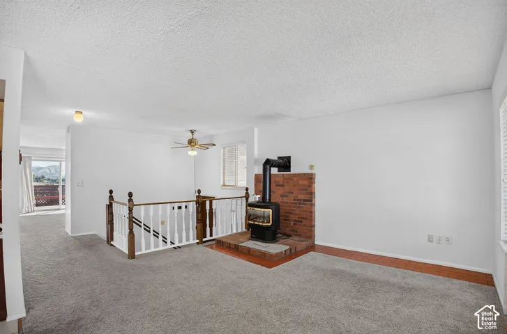 Wood-burning stove with brick surround, carpeted flooring, built-in staircase with wood railing, and ceiling fan with integrated lighting