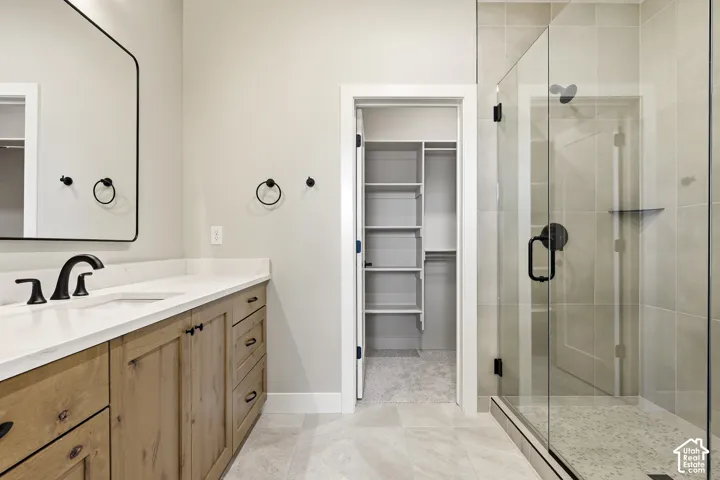 Vanity with a white countertop and natural wood-finish cabinetry, black hardware, frameless shower with tiled walls and a glass enclosure, and a walk-in closet with built-in shelving