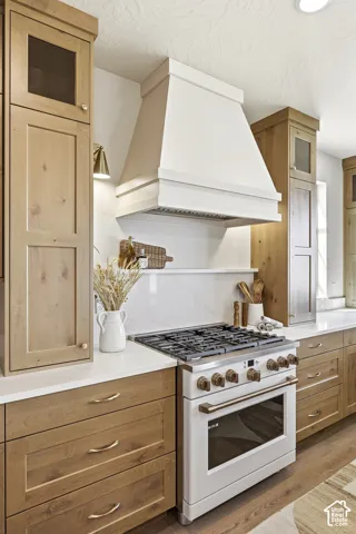 White range hood, natural wood cabinetry, white countertops, built-in gas range, and wood-finish flooring
