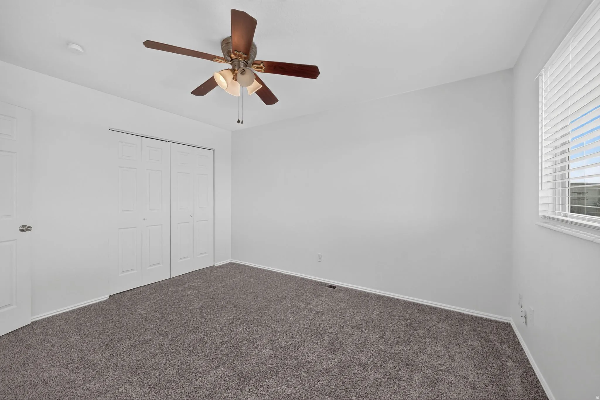 Neutral-toned carpeting with white walls, a multi-pane window with blinds, and a ceiling fan with integrated lighting