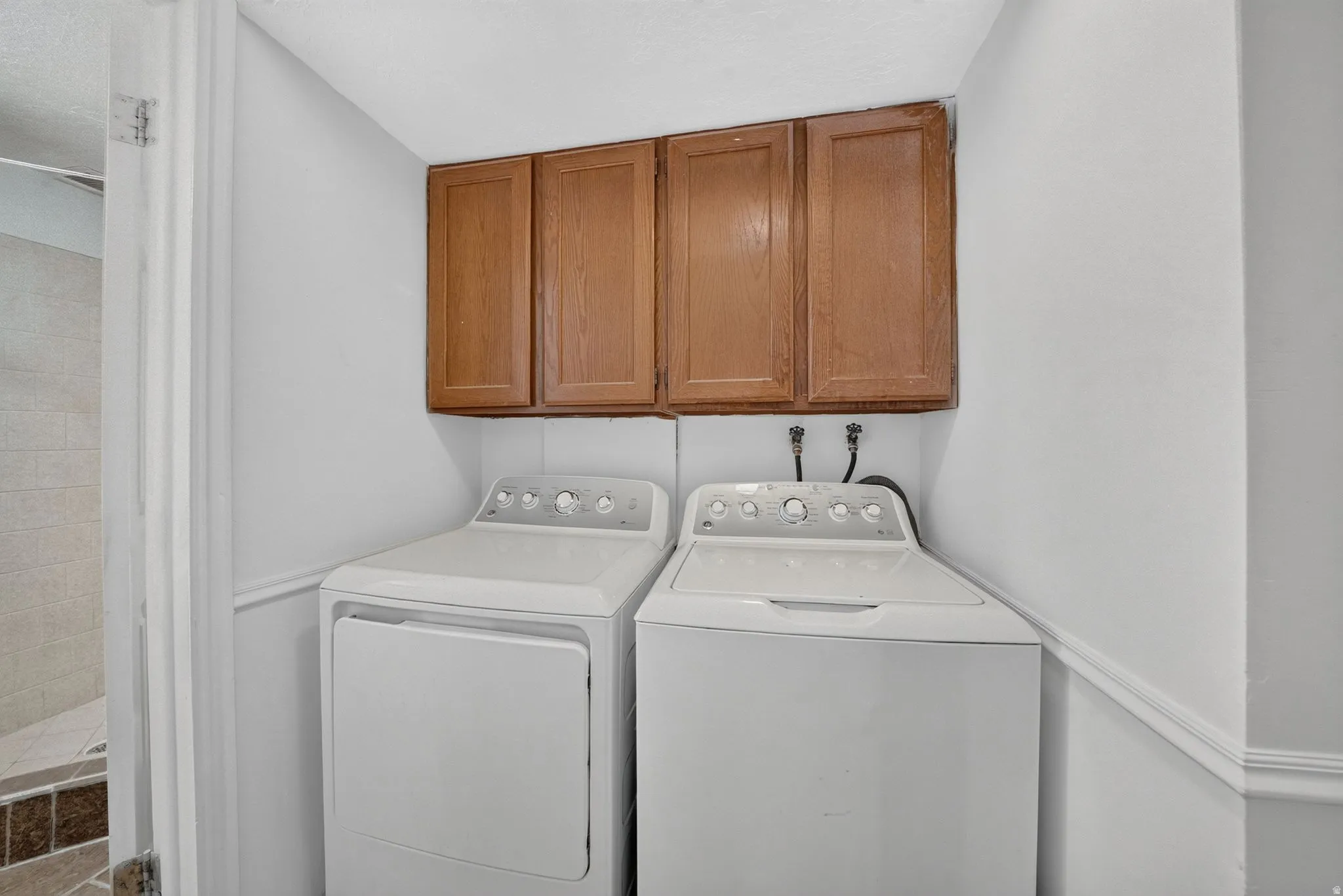 Dedicated laundry room featuring side-by-side white appliances, rich wood-finish cabinetry with ample storage overhead, and bright, neutral finishes throughout