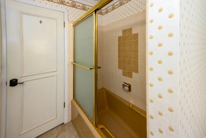 Bathtub with gold-tone sliding glass doors, white subway tile surround with gold-tone accent tiles, and a white paneled door with a black lever handle