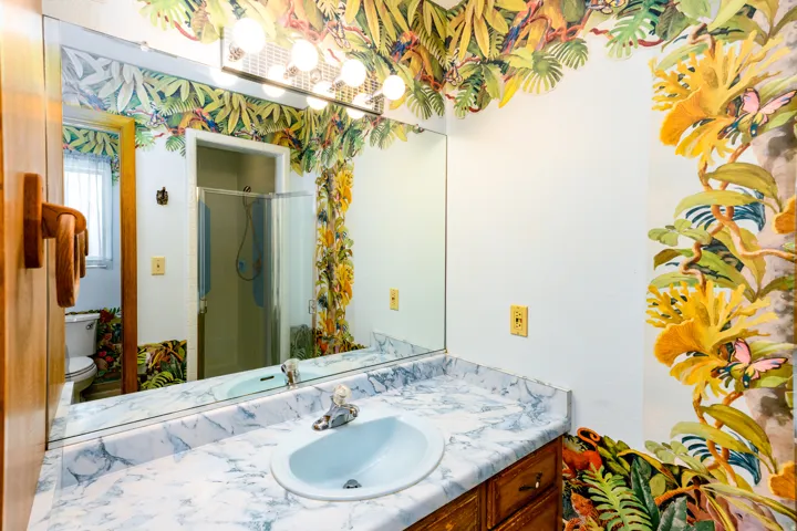 Wall-mounted vanity mirror, single basin sink, white and grey patterned countertop, wood-finish cabinetry, and overhead vanity lighting fixture