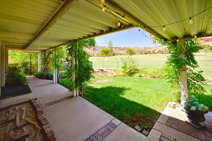 Covered patio with concrete and brick-finish flooring, surrounded by lush greenery, and overlooking a landscaped grassy area