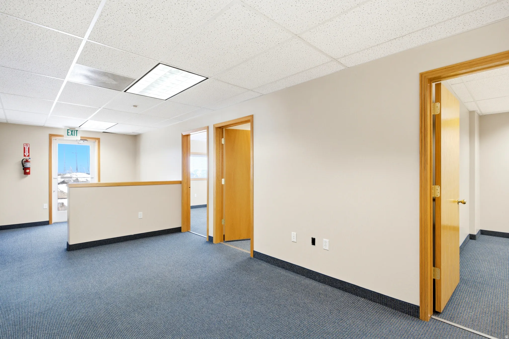 Blue carpet flooring, wood-finish doors, drop ceiling with recessed lighting, half-wall partition with wood-finish railing, and a window with exterior visibility