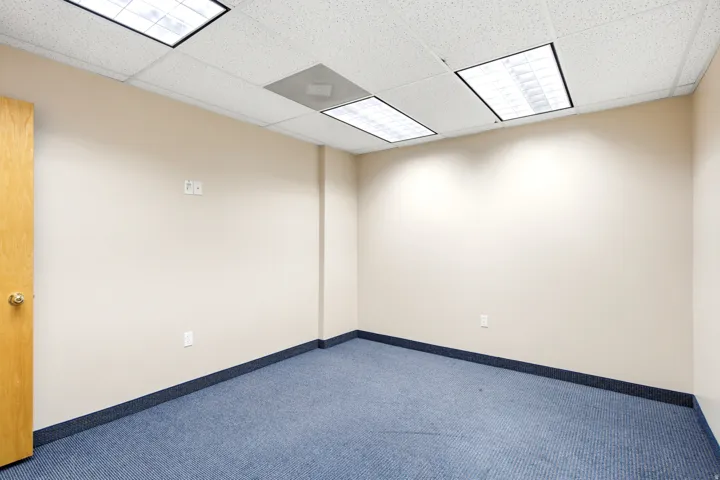 Recessed lighting, drop ceiling, light-toned walls, blue carpet flooring, and a light wood-finish door with a brass-finish knob