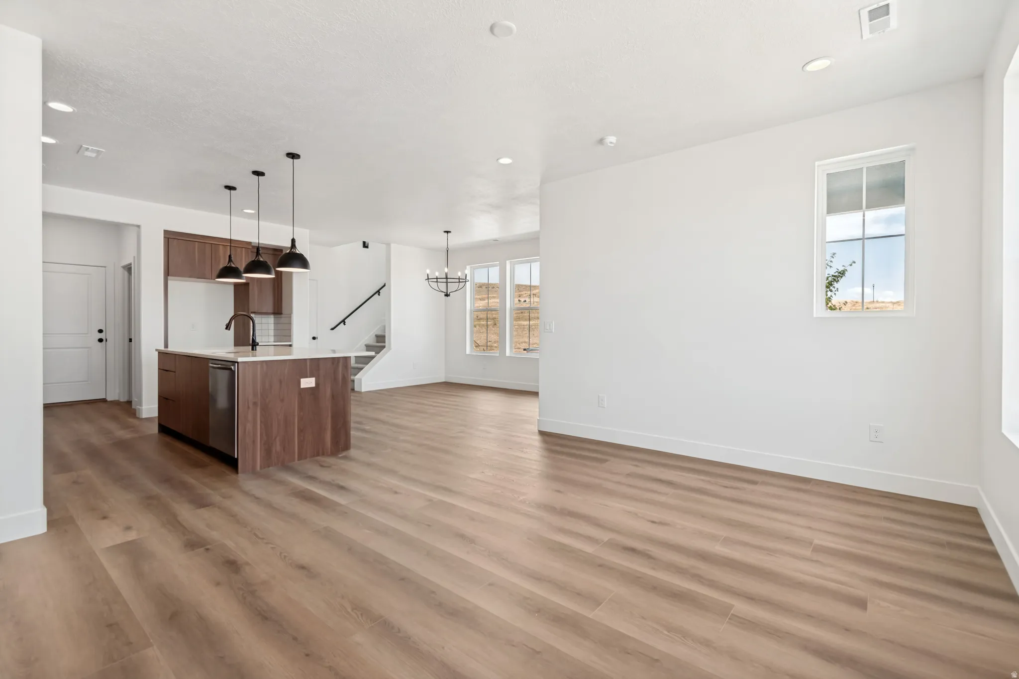 Open-concept living area featuring wood-finish flooring, a kitchen island with integrated sink, modern pendant lighting, and multiple windows for natural illumination