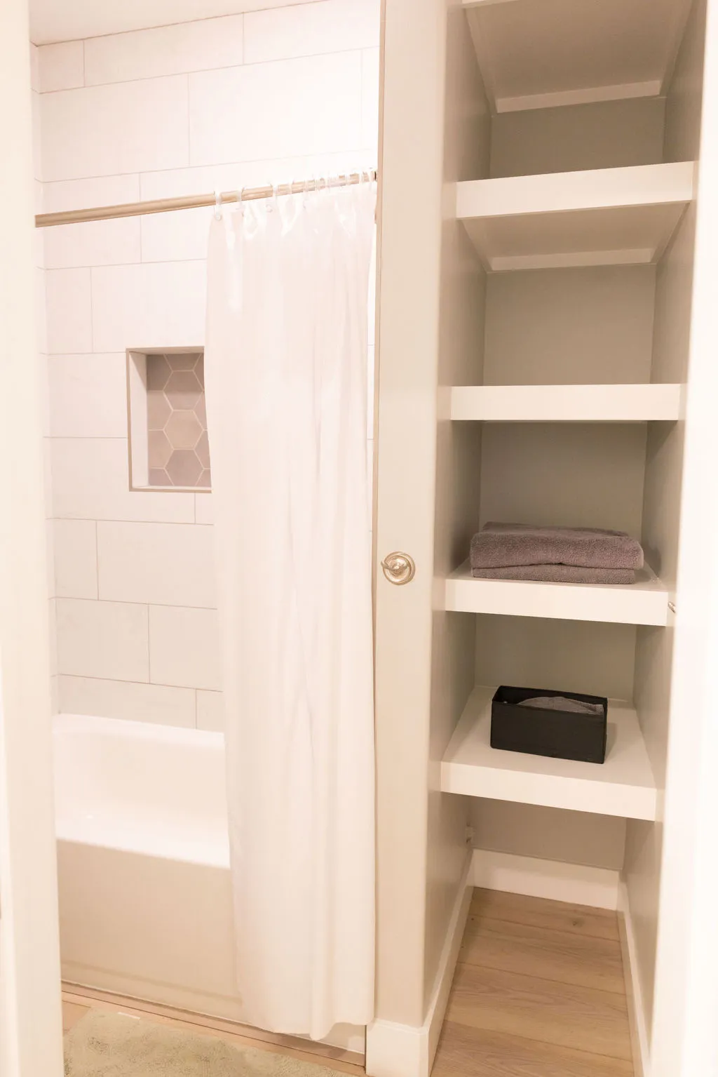 White subway tile shower surround with a built-in hexagonal tile niche, a white soaking tub, and custom built-in shelving with wood-finish flooring
