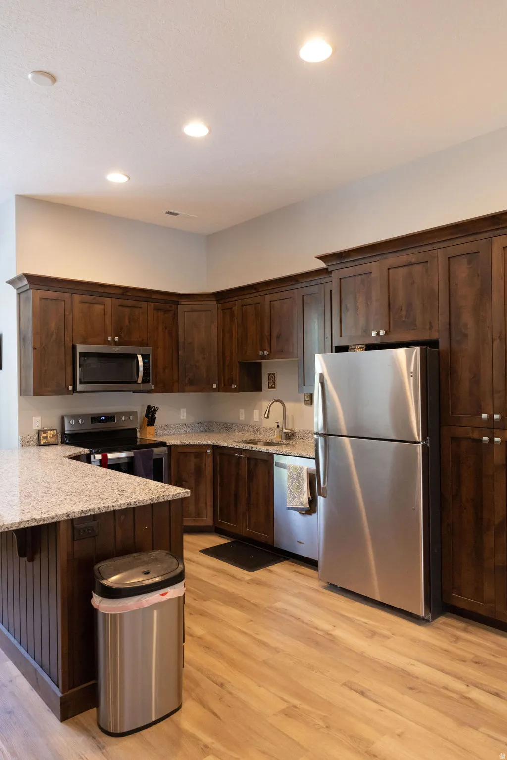Kitchen featuring wood-finish flooring, dark wood cabinetry, stainless steel appliances, speckled countertops, and recessed lighting