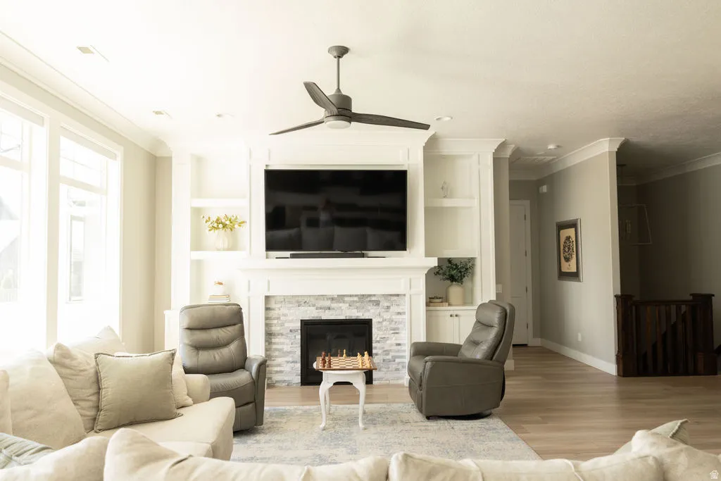 Living area featuring a fireplace with stacked stone surround, built-in shelving, crown molding, a ceiling fan, and wood-finish flooring