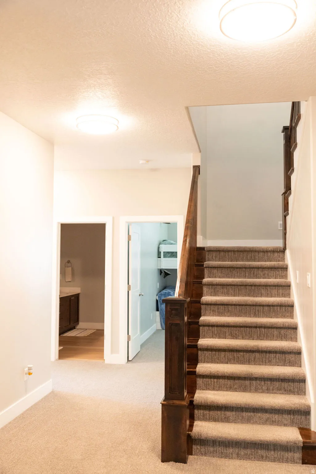 Carpeted staircase with wood newel post and handrail, featuring neutral wall paint, overhead flush mount lighting, and wood-finish flooring visible through an open doorway