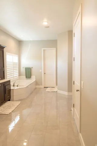 Expansive bath featuring a built-in soaking tub, wood-finish cabinetry, white plantation shutters, white interior doors, and large format polished tile flooring