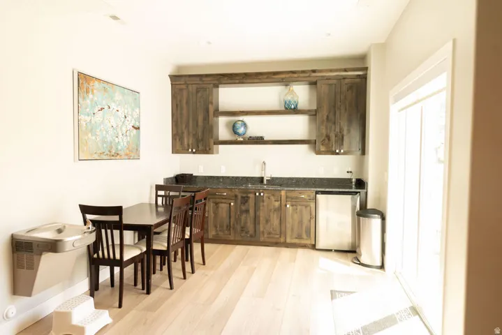 Light-toned wood-finish flooring, built-in dark wood cabinetry, integrated stainless steel beverage refrigerator, dark stone countertops, and a stainless steel sink