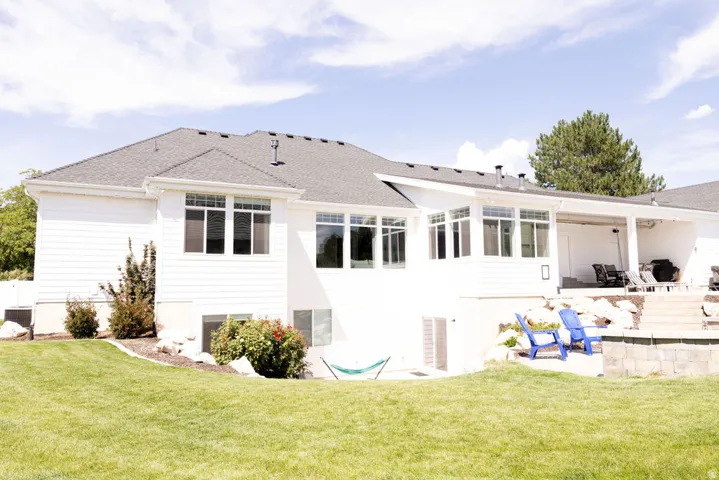 White siding exterior with a dark shingle roof, multiple large windows, covered patio, and a stone retaining wall