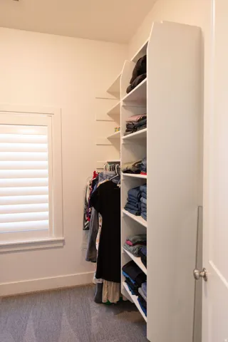 Built-in closet shelving, integrated hanging rod, window with plantation shutters, light-toned wall paint, and gray carpet flooring