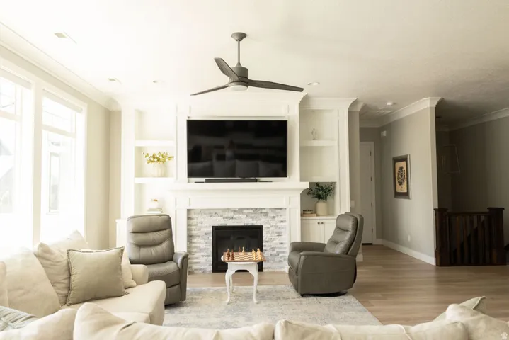 Living area featuring a fireplace with stacked stone surround, built-in shelving, crown molding, a ceiling fan, and wood-finish flooring