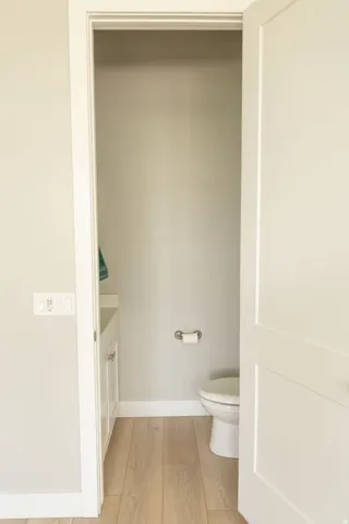 Light wood-finish flooring, white vanity with cabinet storage, white toilet, and white interior door