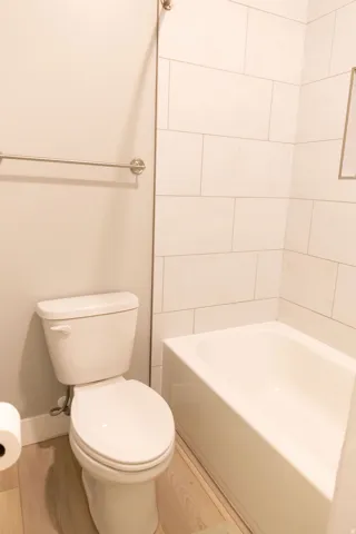 Built-in bathtub with subway tile surround, white toilet, and wood-finish flooring