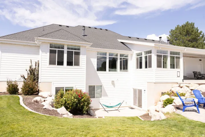 White exterior siding with multiple windows, a dark shingle roof, a concrete patio, and a landscaped yard with a rock feature